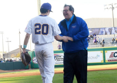 A baseball player in a striped uniform labeled "20" shakes hands with a cheerful older man in a blue jacket on a sunny field.