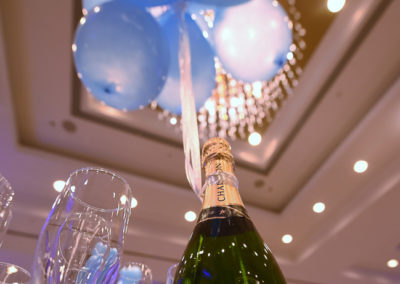 Blue balloons tied to a moët & chandon champagne bottle on a table with glasses, under a chandelier in a banquet hall.