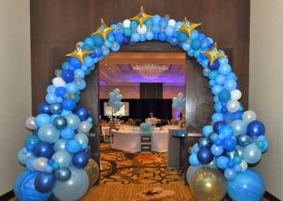 Decorative blue and gold balloon arch at the entrance of a banquet hall with tables set for an event inside.