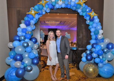 A couple poses under a decorative arch made of blue and gold balloons at a formal event.
