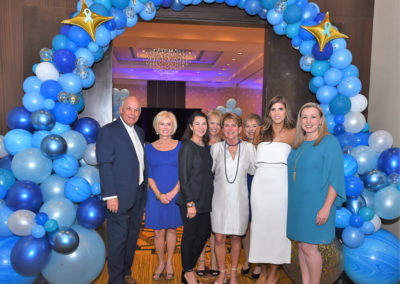 Eight people posing for a photo under a blue and gold balloon arch at an event.