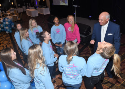 A businessman in a suit laughs and interacts with a group of women in matching pink t-shirts at an indoor event.