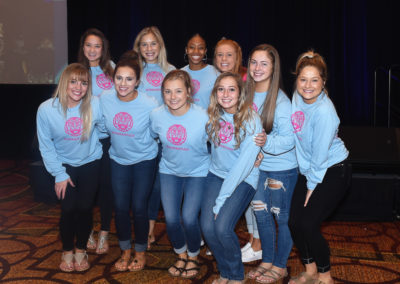 Group of ten smiling women wearing matching light blue t-shirts with pink logos, standing together at an event.