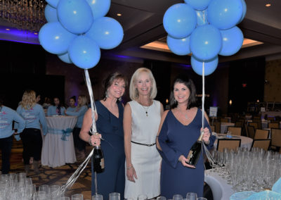 Three women holding blue balloons and smiling at a gala event with a backdrop of glassware and balloon decorations.