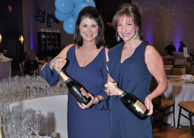 Two women in blue dresses holding champagne bottles at a gala event, standing next to a table with glasses and blue balloons.