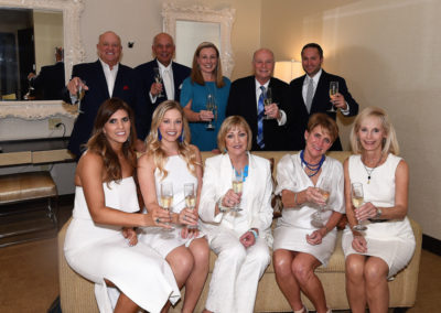 Group of nine adults dressed in formal attire, holding champagne glasses, posing for a photo in an elegant room.