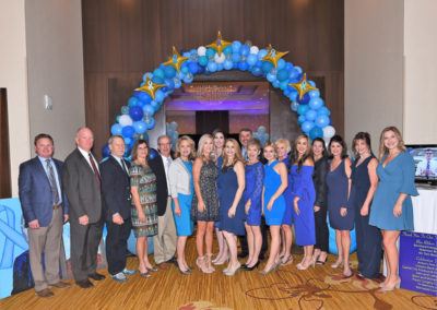 Group of adults standing under a blue and gold balloon arch at a formal event.