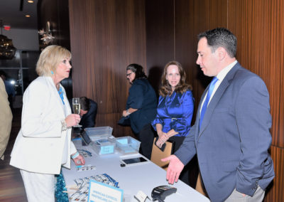 Two people conversing at a registration desk with materials, while a woman in blue observes, in a modern conference room setting.