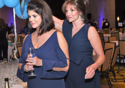 Two women in elegant navy dresses holding balloons at a gala event, one holding a champagne glass, in a festive ballroom setting.