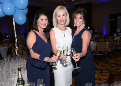 Three women in elegant dresses toast with champagne at a gala event, a table filled with glasses and blue balloons in the background.