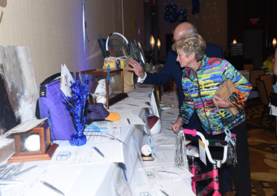 Elderly woman with a walking aid examines items at a charity auction table while interacting with another attendee.