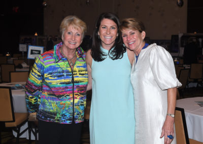Three smiling women pose together at an indoor event, with one woman in a colorful jacket, one in a teal dress, and another in a white dress.