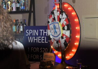 A person stands in front of a light-up "spin the wheel" game at a fundraising event, with a backdrop of a bar.