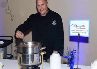 Chef at drusilla seafood restaurant stands with a smile beside a cooking pot and event sign at a catering station.
