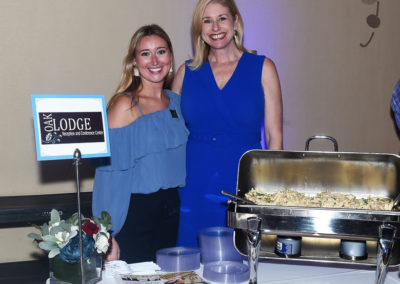Two women smiling at a catering station with a sign labeled "oak lodge" at a formal event.