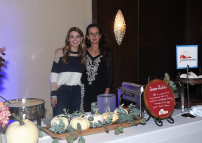 Two women standing next to a buffet table decorated with pumpkins and greenery at an event.