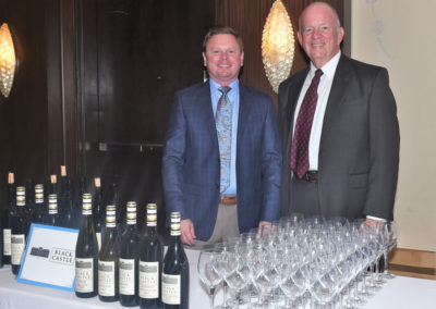 Two men smiling beside a table with wine bottles and empty glasses at an event.