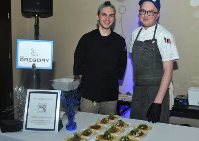 Two chefs standing at a food station with a sign labeled "gregory" presenting dishes of bruschetta at an event.