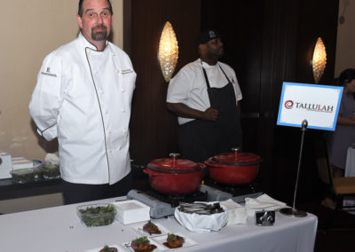 Two chefs standing by a buffet setup with serving dishes and small plated appetizers, featuring a sign that reads "tallulah.