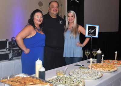 Three people standing behind a food table at a catering event, smiling at the camera.