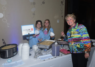 Three women smiling at a catering station with food warmers and a roux 61 sign at a festive event.