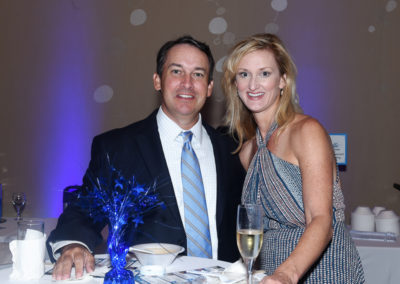A man in a suit and a woman in a striped dress smiling at a table with champagne and decorative blue centerpiece at an event.