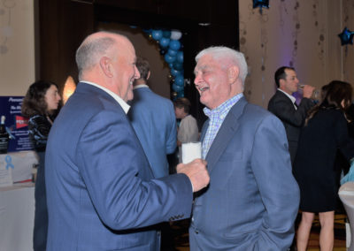 Two elderly men in suits chatting and smiling at a formal event with a balloon-decorated background.