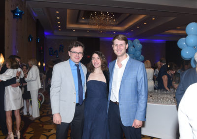 Three people smiling at a party with blue balloons and a chandelier in the background.