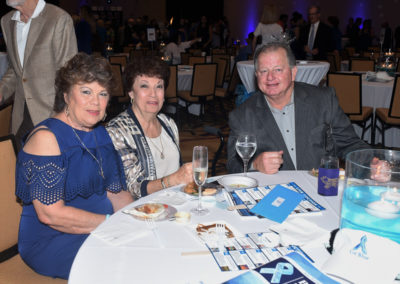 Three mature adults seated at a banquet table with dinner plates and drinks during an event, smiling at the camera.