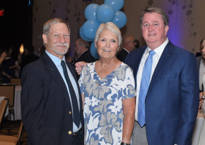 Three senior adults standing together and smiling at a formal event with blue balloons in the background.