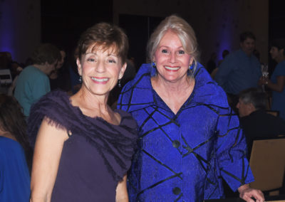 Two smiling older women standing together at a social event, one in a purple dress and the other in a blue coat, with a wine glass in hand.