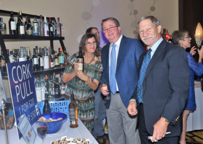 Two men and a woman standing by a "cork pull" station with bottles and desserts during an event.