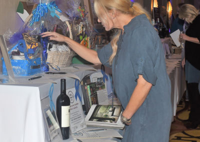 Woman in a blue dress examining items at a charity auction table with baskets and wine bottles.