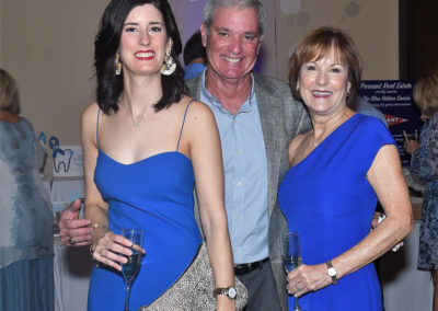 Two women and a man in formal wear, smiling, holding glasses at an event; one in a blue dress, one in a blue strapless dress, and the man in a casual shirt.