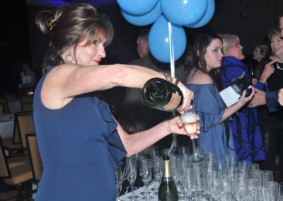 A woman in a navy blue dress pours champagne into glasses at a festive event, surrounded by people and blue balloons.