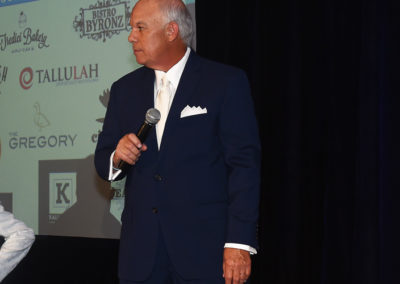 An older man in a blue suit and white pocket square holding a microphone, standing next to a projector screen displaying logos.