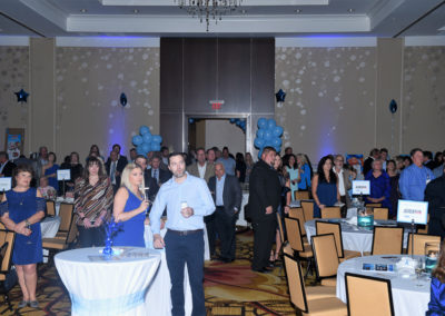 People standing and sitting in a banquet hall decorated with blue balloons for an event.