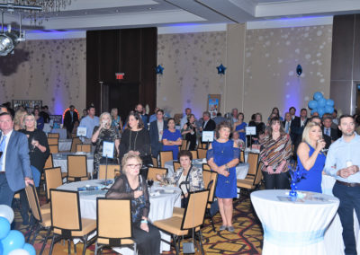 A group of people attentively watching a presentation at a gala event decorated with blue balloons and tables.