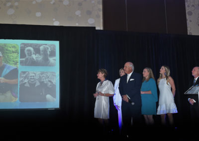 A group of people stand on stage at an event, looking at a large screen displaying various personal photographs.