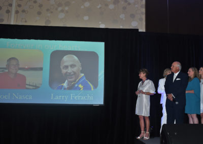 People on stage at an event beside a screen displaying tributes to individuals named joel nasca and larry ferachi.