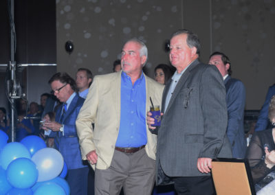 Two middle-aged men in business casual attire standing at a social event with blue balloons and a crowd in the background.