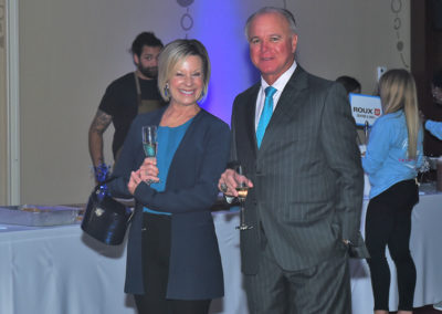A man and a woman holding wine glasses and smiling at a formal event, standing in a room with blue themed decorations.