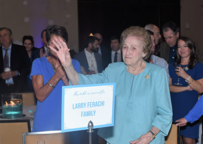 Elderly woman in a blue suit standing at a table with a "larry ferachi family" sign, clapping her hands at an event.