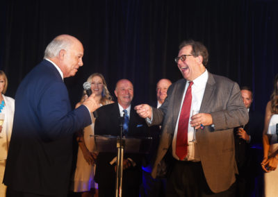 Two men laughing and talking at a podium during a formal event, with people in the background and a spotlight shining on them.
