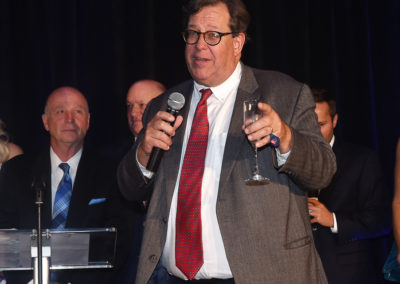 A man in a suit with glasses holding a microphone and a glass of wine, speaking at a podium during an event.