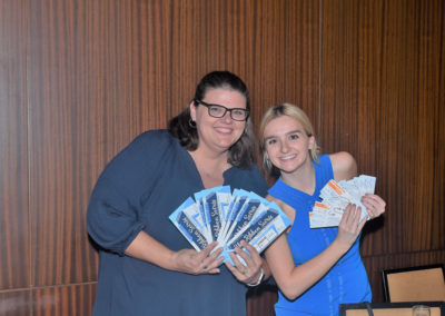 Two women smiling and holding multiple tickets, standing in front of a wood-paneled wall at an event.