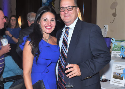 A man and woman in formal attire smile for the camera at an event, the woman in a blue dress and the man holding a wine glass.