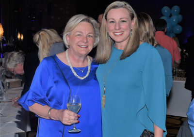 Two women smiling at a formal event, one holding a wine glass, both dressed in elegant blue outfits with necklaces.