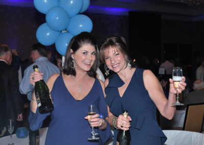 Two women in blue dresses holding champagne glasses and a bottle, with blue balloons in the background at a festive event.