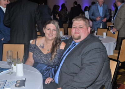 A couple dressed in semi-formal attire sitting at a table during a gala event, with other guests mingling in the background.